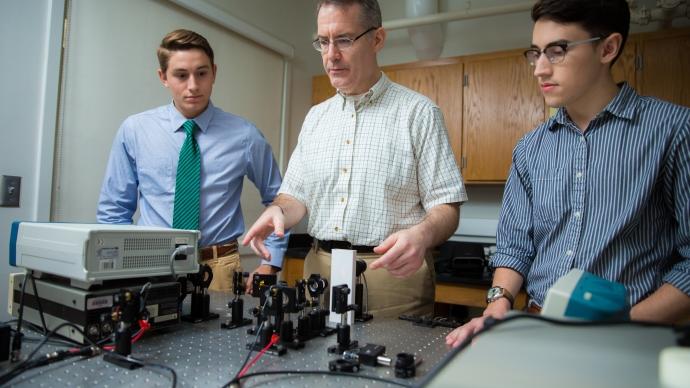 Dennis Ugolini stands between Sean Farrell and Matt Jenkins behind a table full of small-scale LIGO equipment
